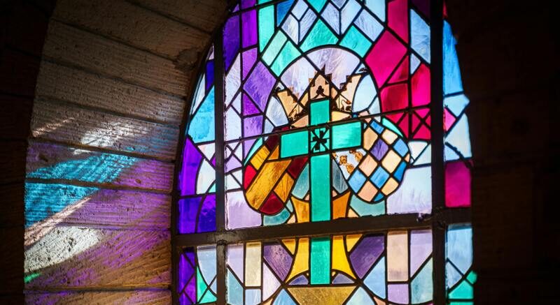 Vibrant stained glass window with a central cross motif casting multicolored light onto a stone interior — church pew refurbishing, stained glass windows.