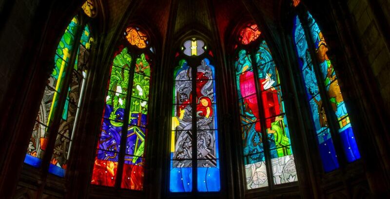 Tall arched stained‑glass windows with vivid multicolored panels depicting religious motifs, casting colored light into a dark church interior — church pew refurbishing, stained glass windows.